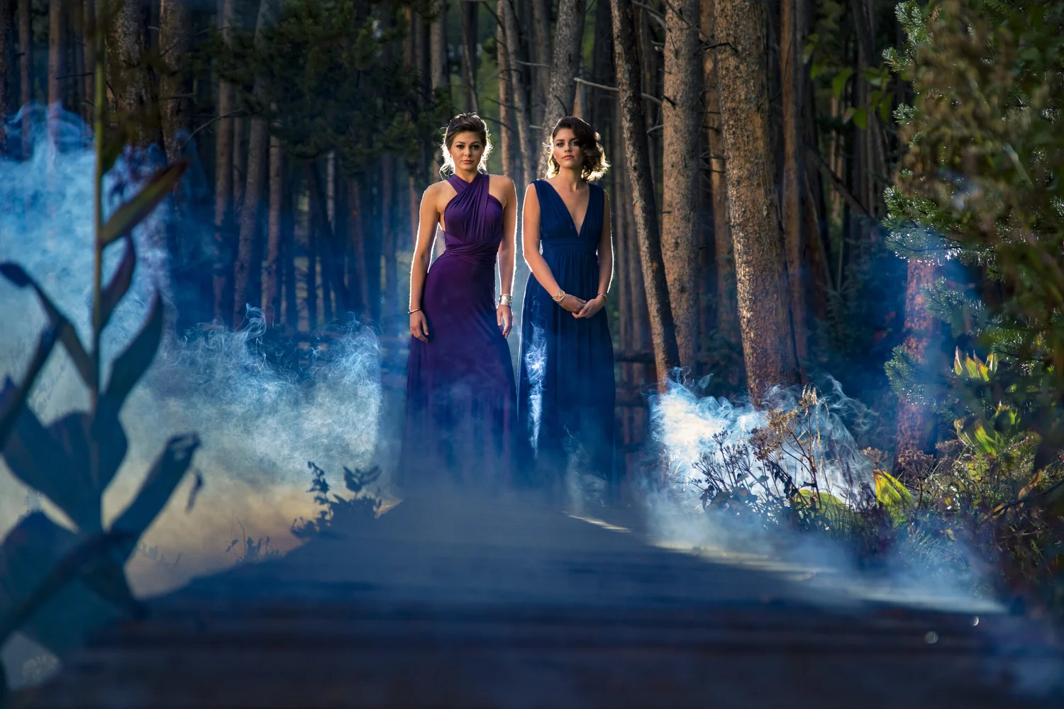 Two women in formal wardrobe staged inside a cinematic smoke-filled forest