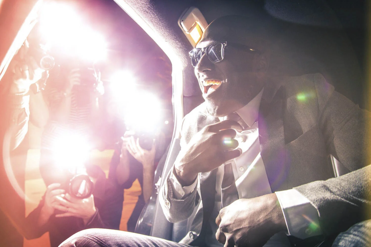 Man in sunglasses photographed inside a vehicle amid press camera flashes
